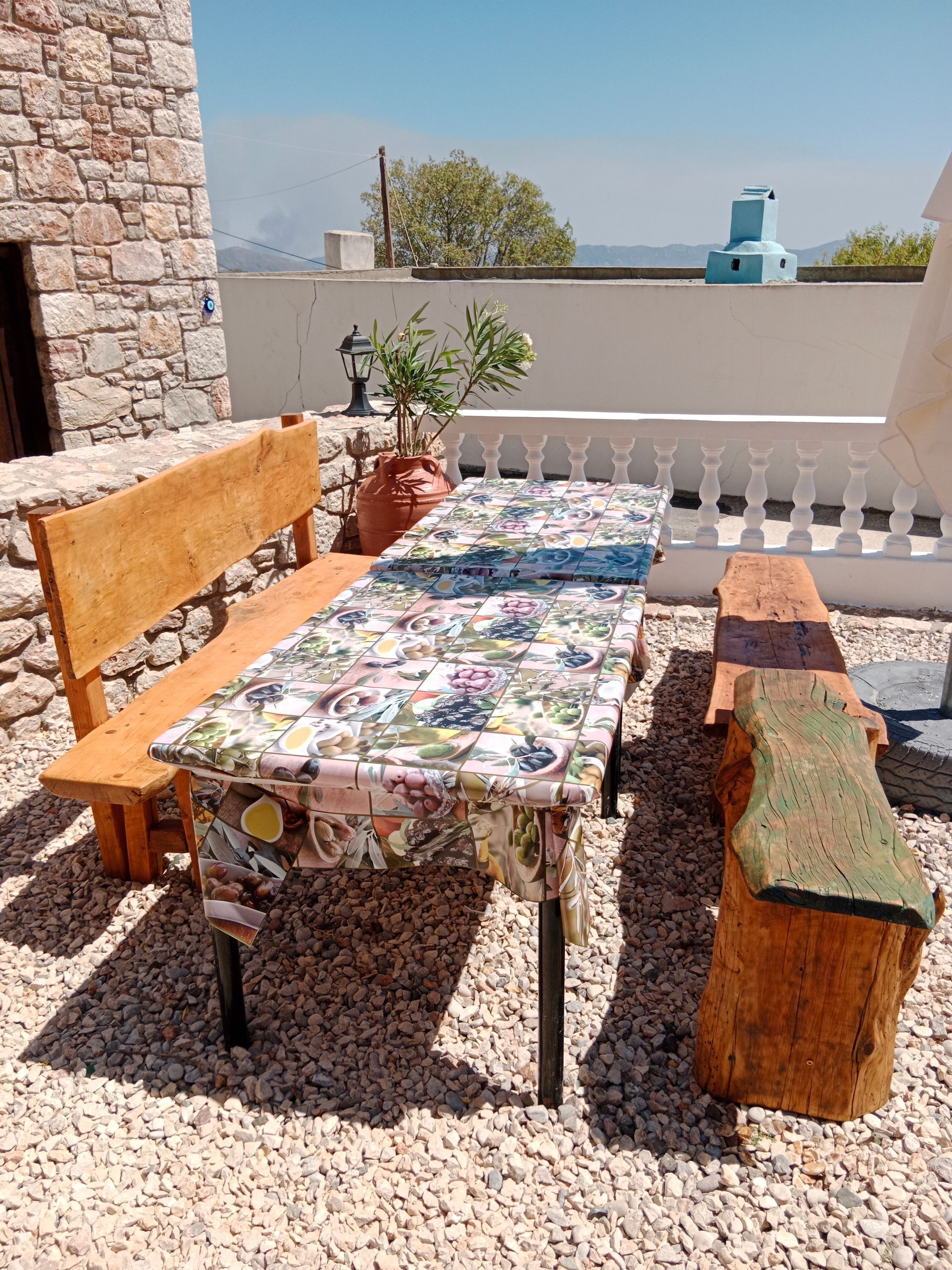 An outdoor seating area with a wooden bench, a table covered with a colorful tablecloth, and a potted plant.