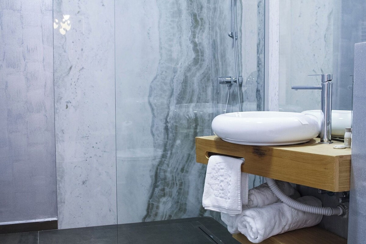 A bathroom with a marble wall, a wooden vanity, a white sink, a shower, and a towel rack.