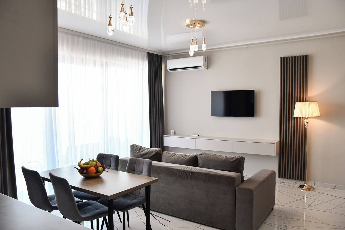 A modern living room with a sofa, dining table, and chairs. There is a flat-screen TV mounted on the wall, a floor lamp, and an air conditioning unit.