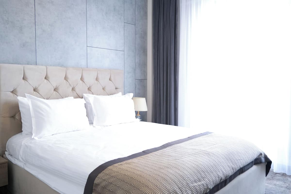 A hotel room with a tufted headboard, a bed with white bedding, a patterned throw blanket, a bedside lamp, and a view of the outdoors through a window with curtains.