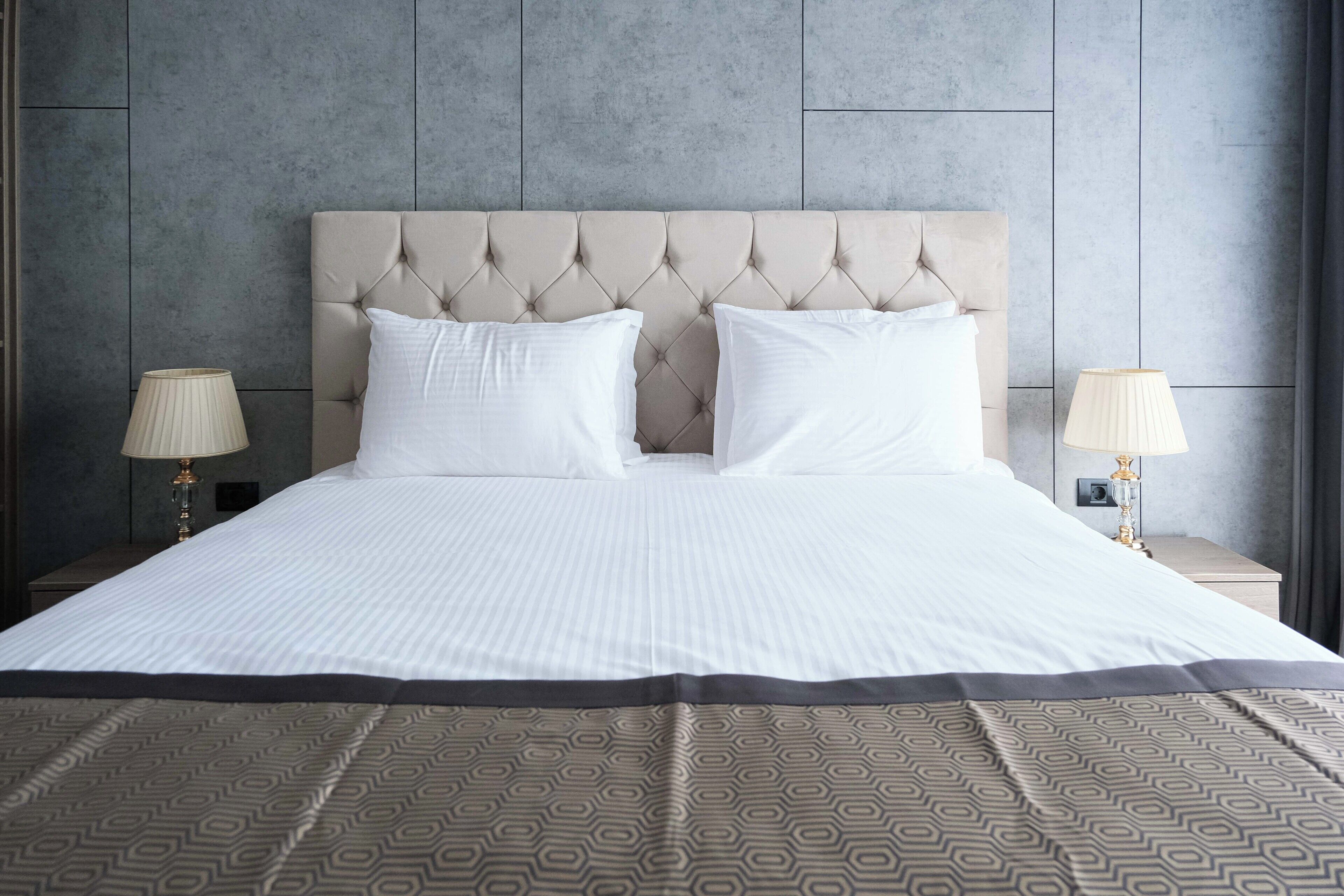 Una cama bien hecha con cabecera acolchada, dos lámparas de noche y una colcha estampada.