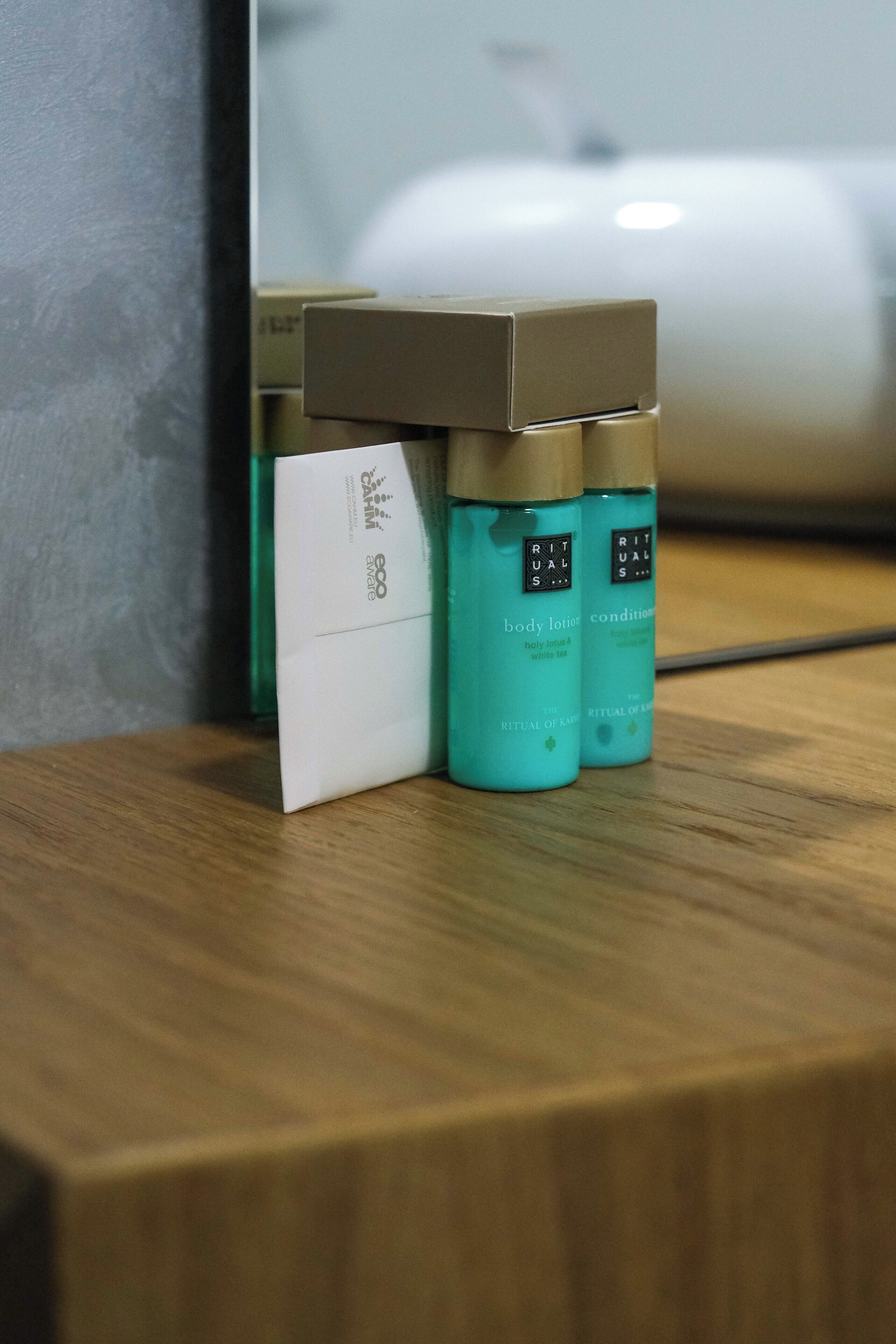 A wooden surface with a white envelope and two teal bottles labeled "Rituals" on a reflective surface.