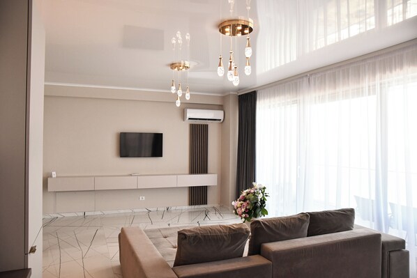 A modern living room with a sofa, a flat-screen TV mounted on the wall, a decorative chandelier, and a large window with sheer curtains.