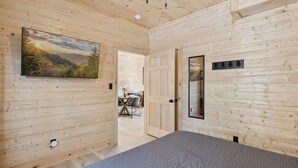 A room with wooden walls, a bed, a painting, and a door leading to another room. - Black Bear by Beyond Expectations: 2BR Cabin w/ Hot Tub, Arcade, & Axe Throwing (Sevierville)