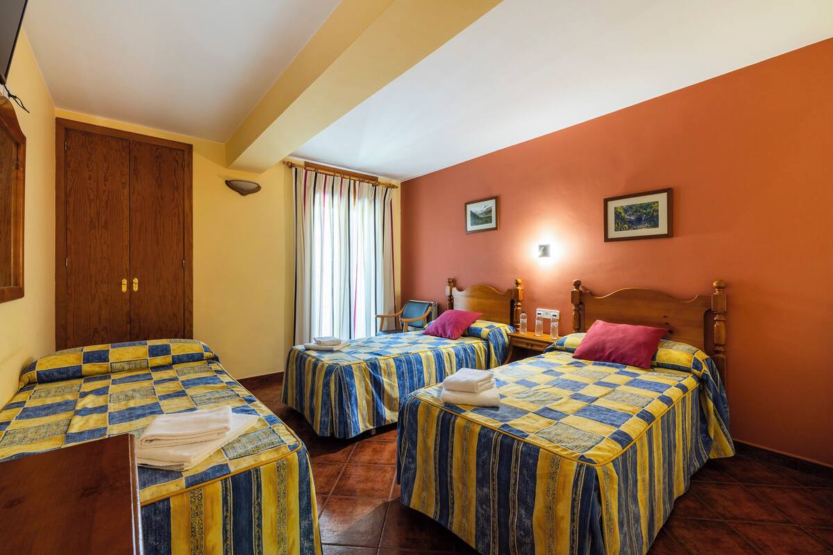 A hotel room with two beds, wooden headboards, a wooden wardrobe, a chair, and a window with curtains.