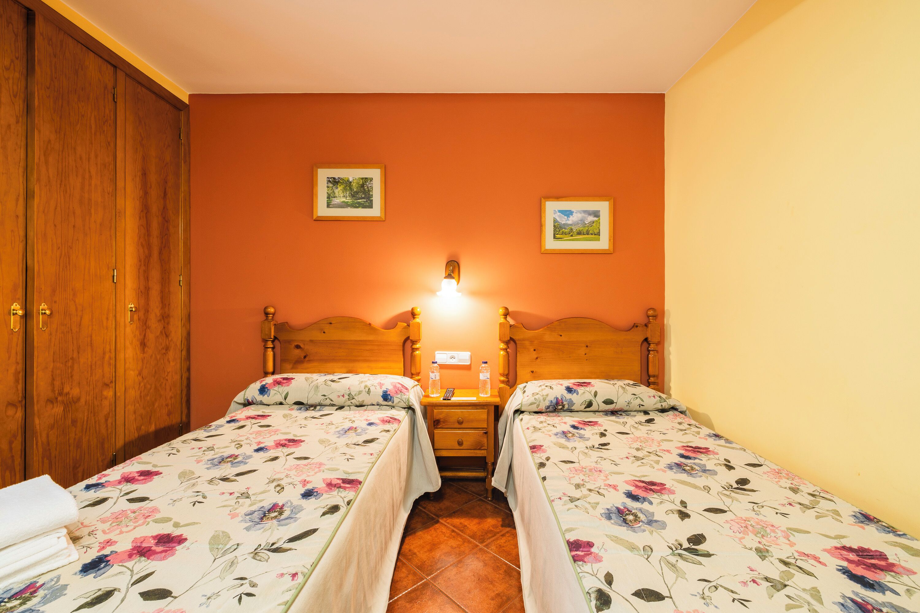 Une chambre avec deux lits simples, des têtes de lit en bois, une table de chevet avec une lampe et deux tableaux encadrés au mur.