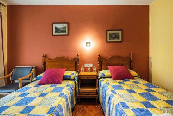 Habitación de hotel con dos camas, una mesita de noche con lámpara, una silla y dos cuadros en la pared.