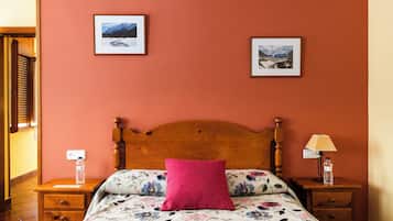 Een slaapkamer met een houten hoofdbord, bloemenbedrukking, een roze kussen, twee ingelijste foto’s aan de muur, een nachtkastje met een lamp en een waterfles.