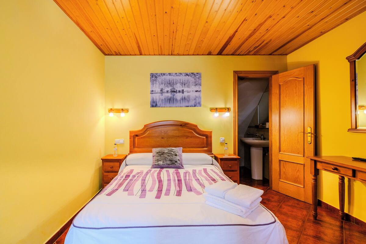 A bedroom with a wooden ceiling, a bed with a white and purple bedspread, two bedside lamps, a nightstand, a wooden wardrobe, and a bathroom visible through an open door.