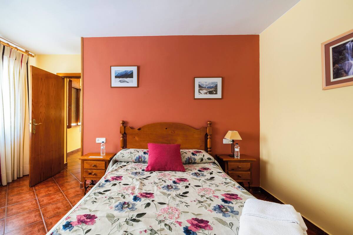 A bedroom with a wooden bed, a floral bedspread, a pink pillow, two framed pictures on the wall, and a nightstand with a lamp.