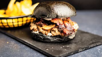 Hamburguesa con tocino y champiñones en un pan negro, acompañada de papas fritas sobre una bandeja negra.