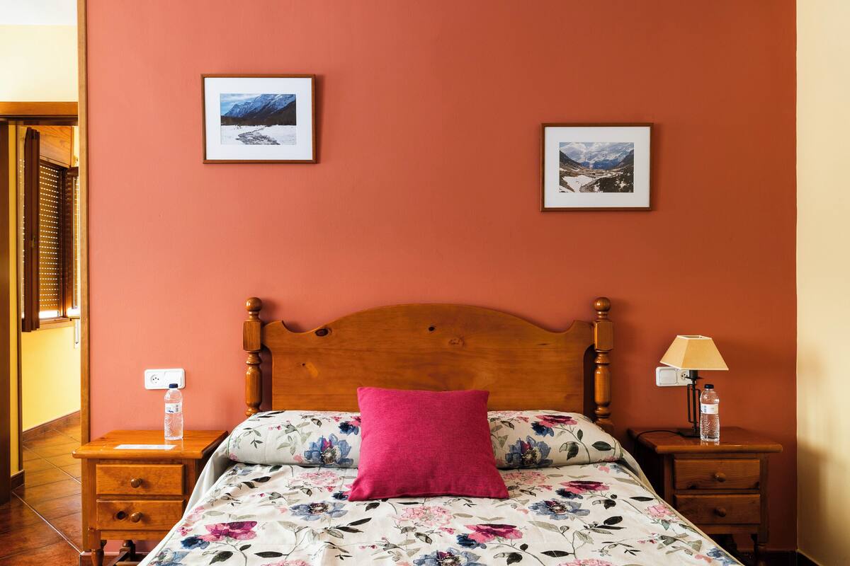 A bedroom with a wooden headboard, floral bedding, a pink pillow, two framed pictures on the wall, a bedside table with a lamp, and a water bottle.