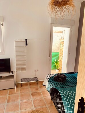 A bedroom with a bed, a TV, a shelf, a small table, and a window with a view of a garden. - Apartment Tequila, City Center Studio Apartment (Almunecar)