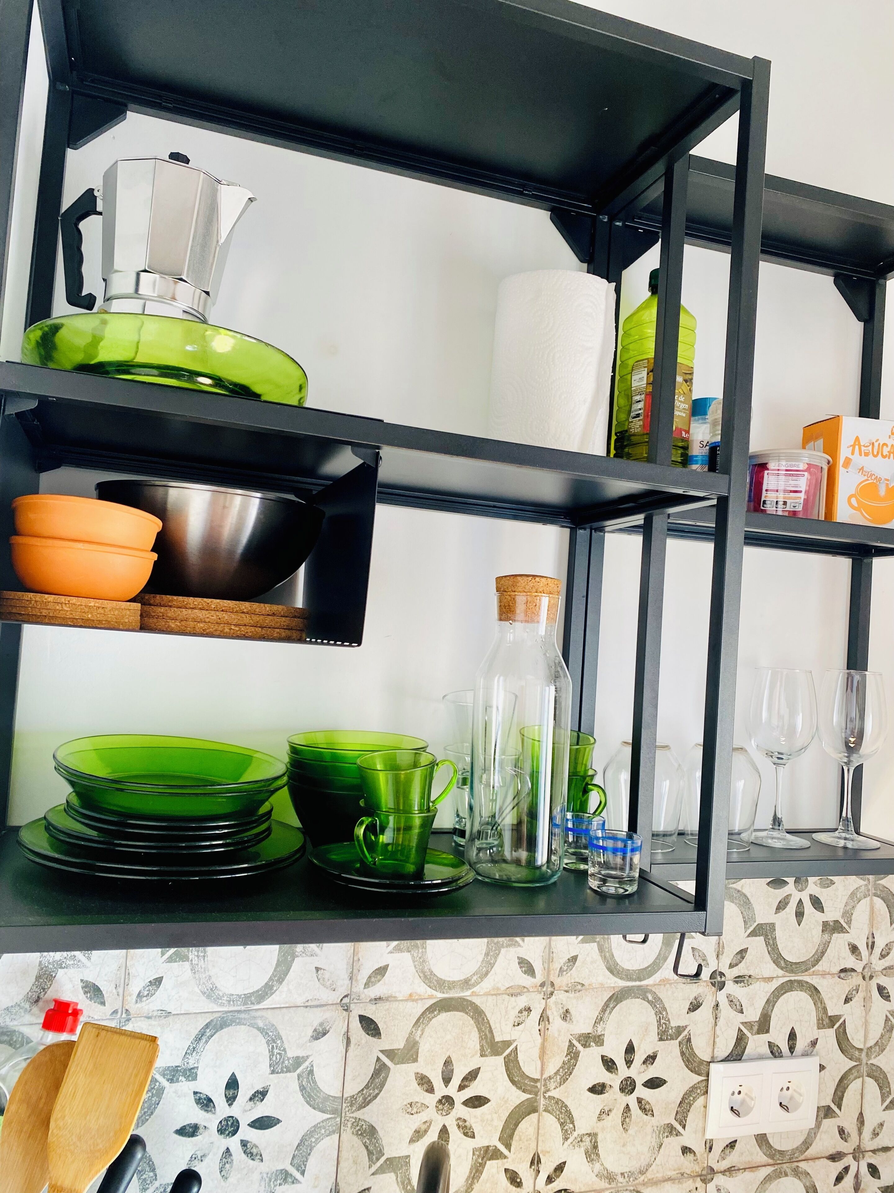 A kitchen with black metal shelves holding various kitchenware, including green and orange bowls, a stainless steel pot, and a green kettle.