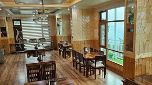 A spacious dining area with wooden tables and chairs, large windows, and a chandelier. - Hotel Sarahan Inn (Rampur)