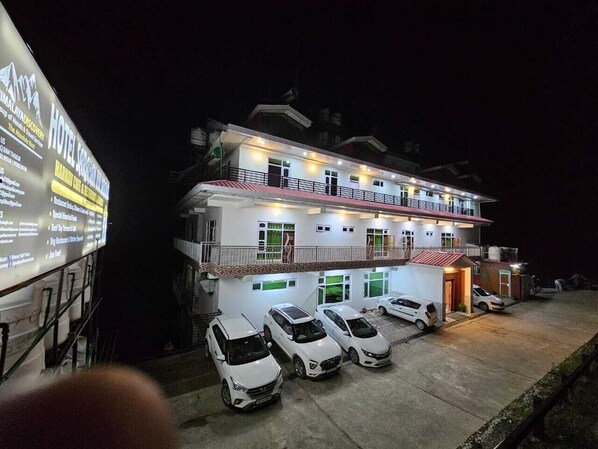 A two-story hotel with a balcony on the upper floor, multiple cars parked in front, and a large illuminated signboard to the left.