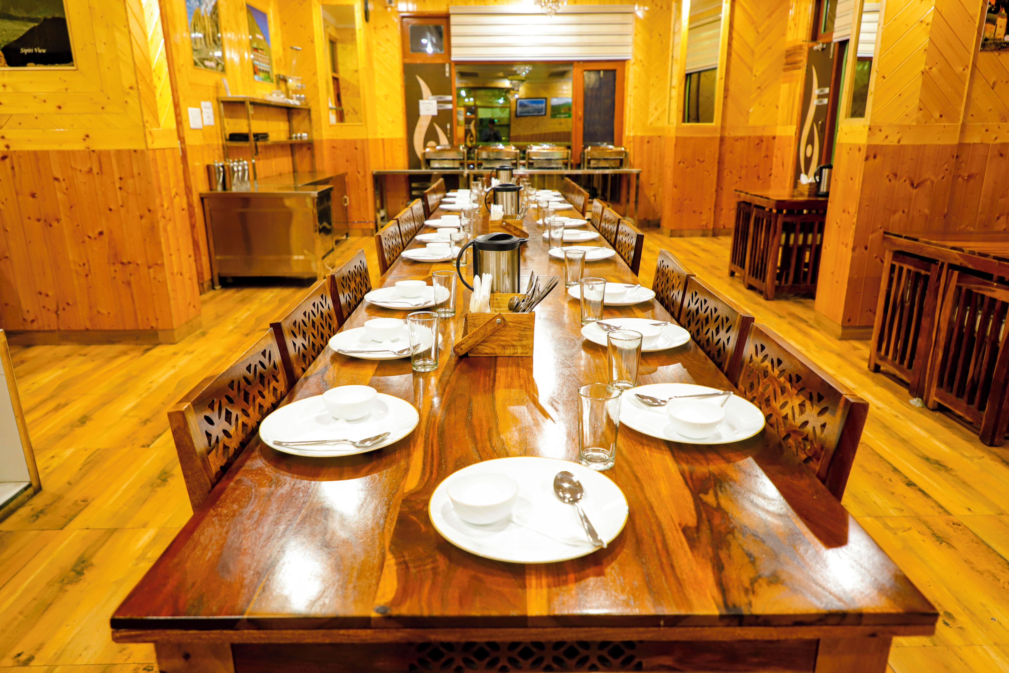 A long wooden dining table set for a meal with chairs on both sides, a buffet area in the background, and a kitchen visible through an open door.