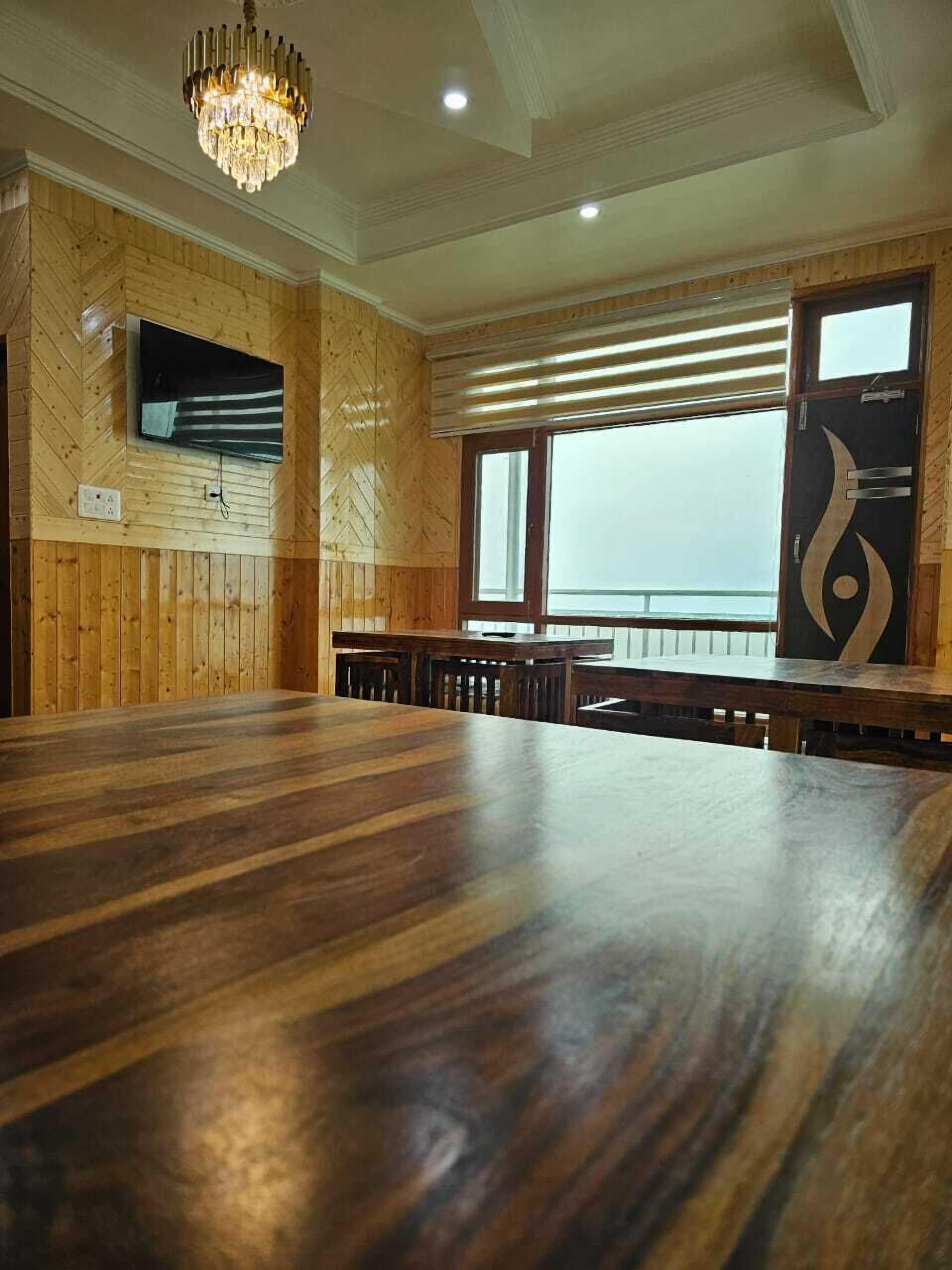 A spacious room with a large wooden dining table, a chandelier, a television mounted on the wall, and a window with blinds.