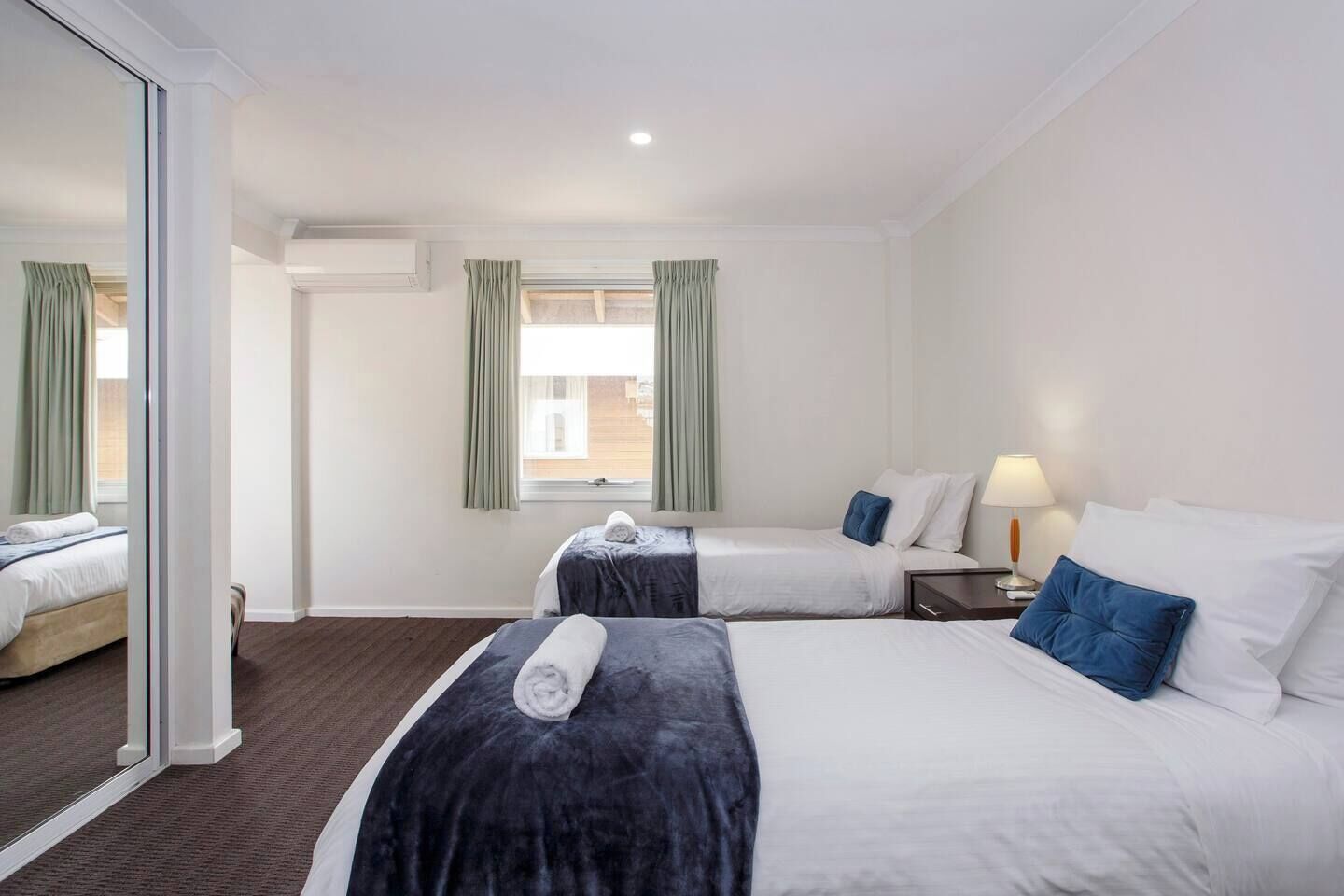 A hotel room with two beds, a large mirror, a window with curtains, a bedside table with a lamp, and an air conditioning unit.