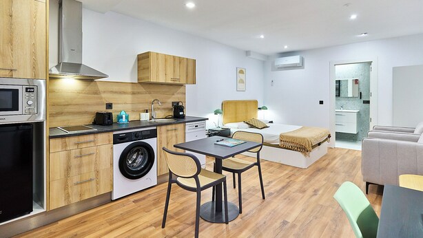 Una cocina moderna con microondas, lavadora y armarios de madera. Hay una zona de comedor con una mesa y sillas, y una cama al fondo.