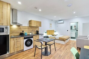 A modern kitchen with a microwave, washing machine, and wooden cabinets. There is a dining area with a table and chairs, and a bed in the background. - Apartamentos Beatriz by gaiarooms (Salamanca)