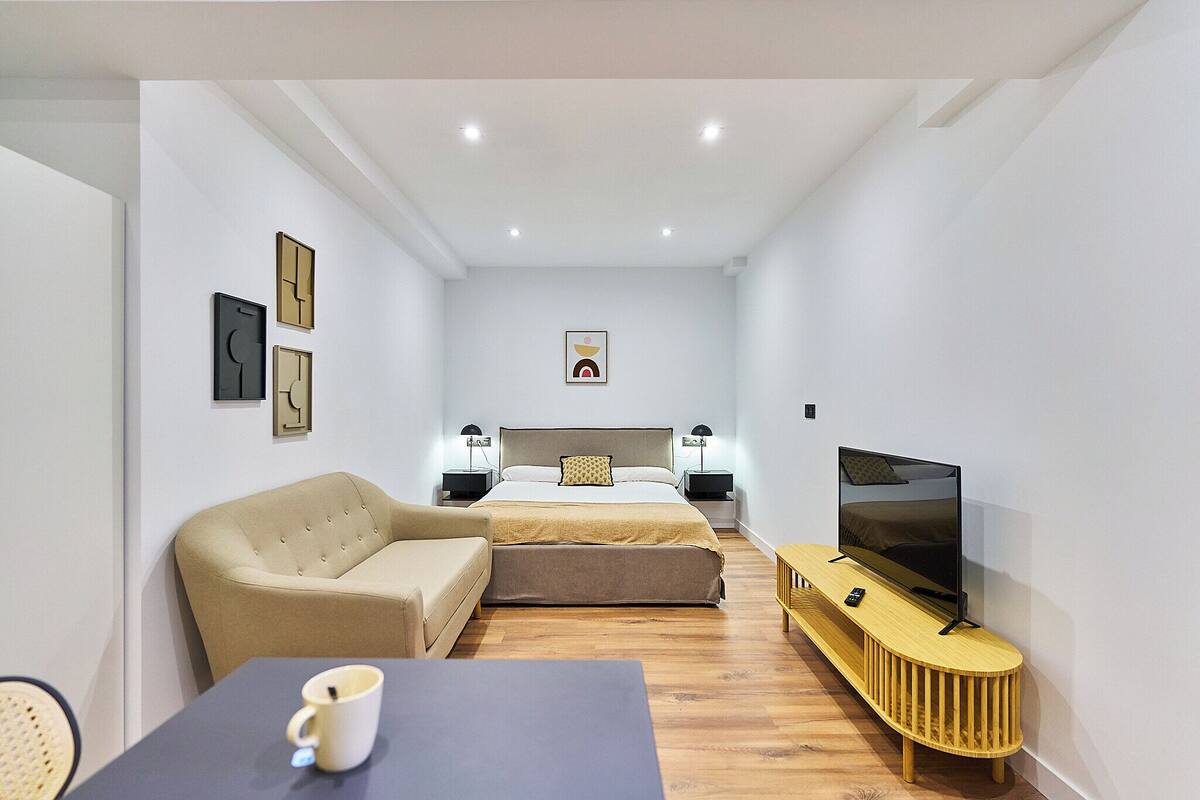 A modern hotel room with a bed, a sofa, a TV, and a small table with a cup.