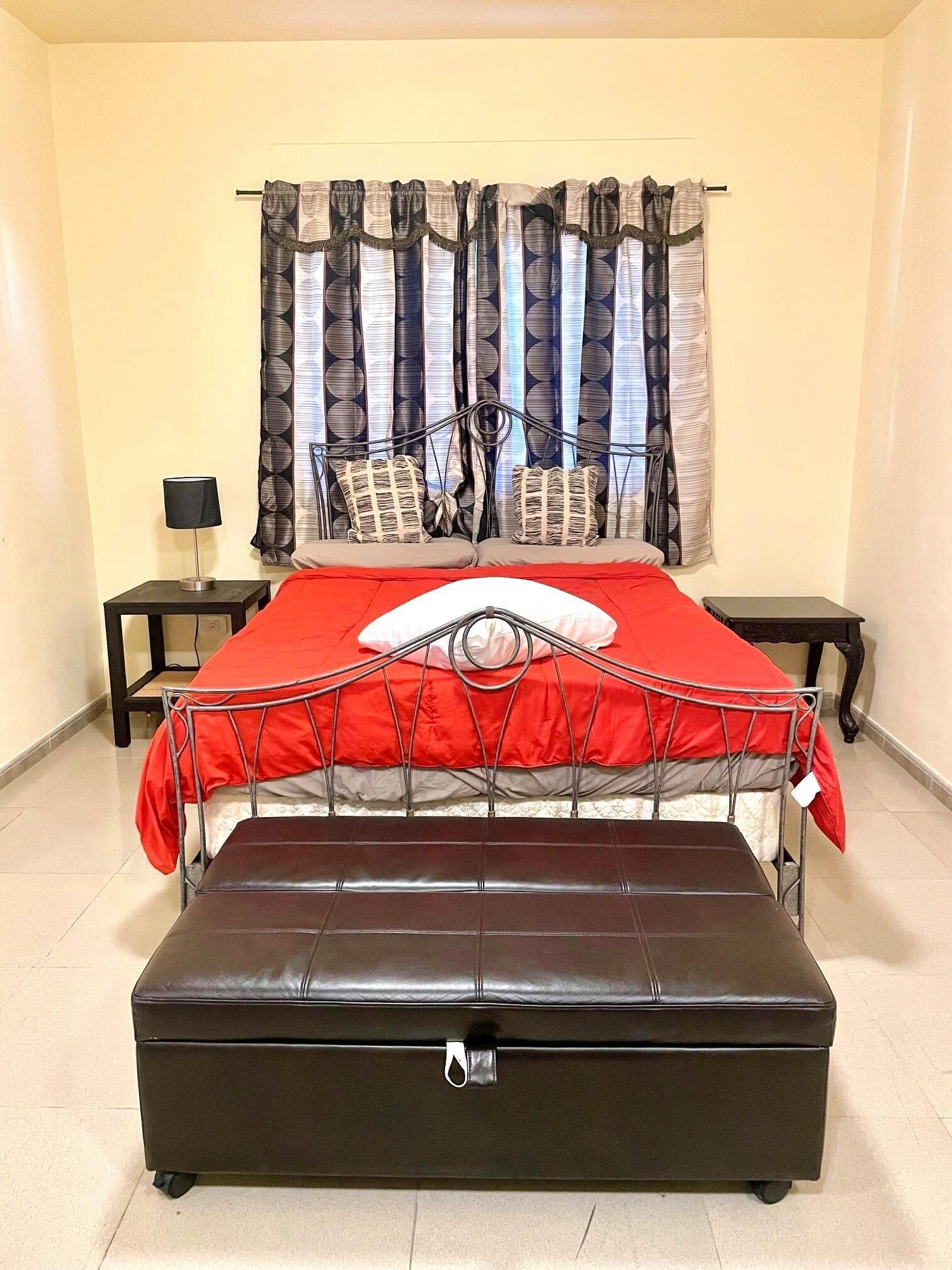 A bed with a metal frame, a red and white bedspread, and a black leather bench. There are two bedside tables with lamps.