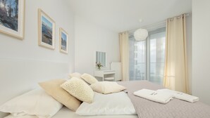 Een net opgemaakt bed met kussens, een nachtkastje met een vaas en een raam met gordijnen.