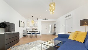Een moderne woonkamer met een blauwe bank, een glazen salontafel en een donkere dressoir. Er is een keuken zichtbaar op de achtergrond met witte kasten en een eethoek.