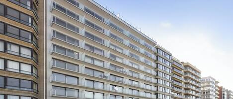 A multi-story residential building with balconies, a parking area, and a clear sky.