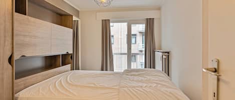 Une chambre à coucher avec un lit, des étagères en bois intégrées, un luminaire suspendu et une vue sur les bâtiments par une fenêtre.