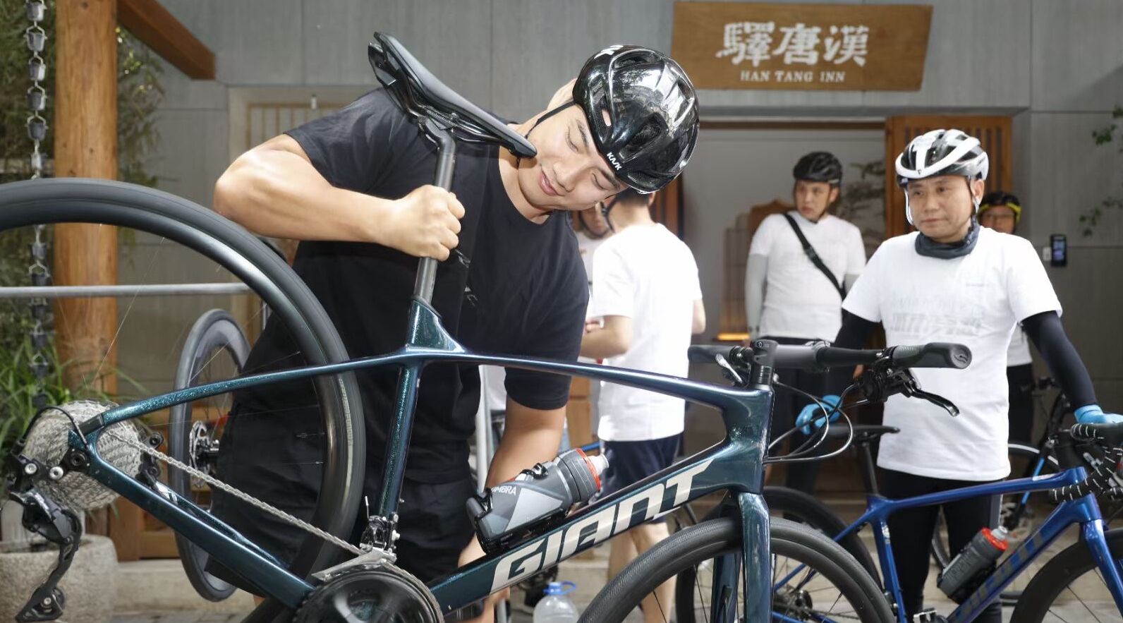 Una persona sistema una bicicletta con telaio del marchio 'GIANT', un'altra indossa una camicia bianca e una terza persona, anch'essa con una camicia bianca e un casco, davanti a un edificio con un cartello che riporta la scritta 'HAN TANG INN'.