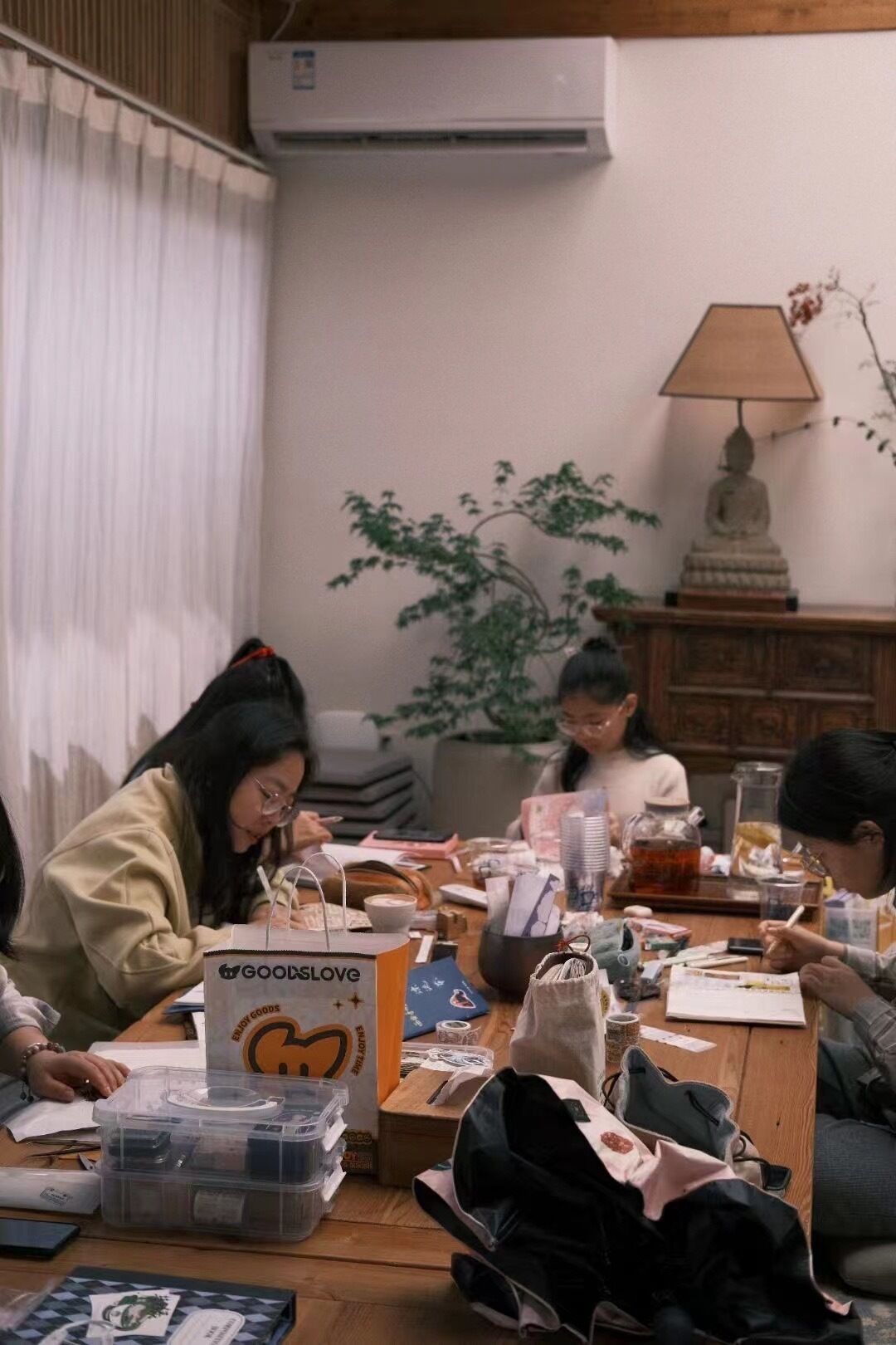 A group of people seated around a wooden table in a room with a white air conditioner, a lamp, and a plant.