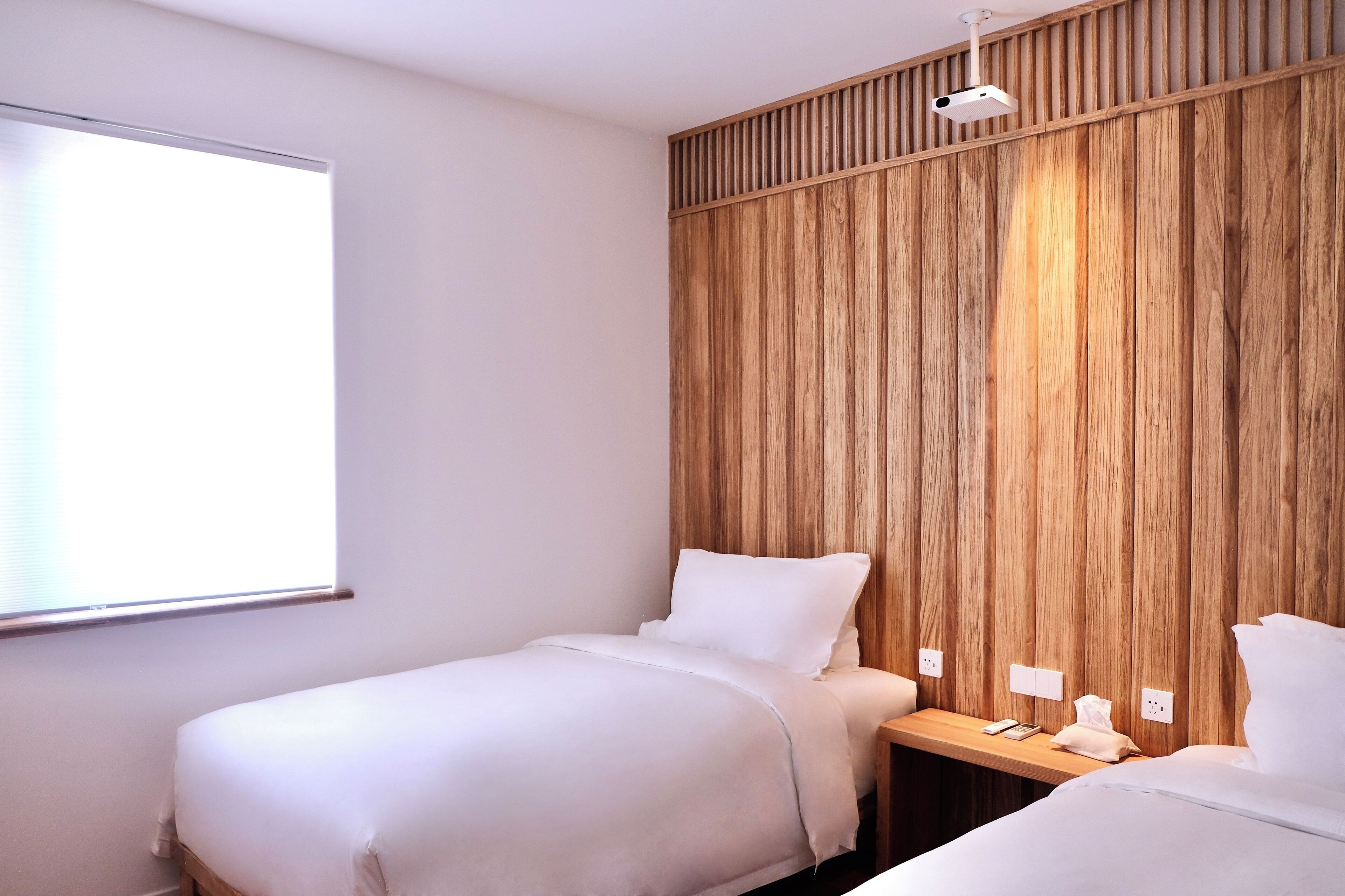A hotel room with two beds, a wooden panel wall, a projector, and a window.