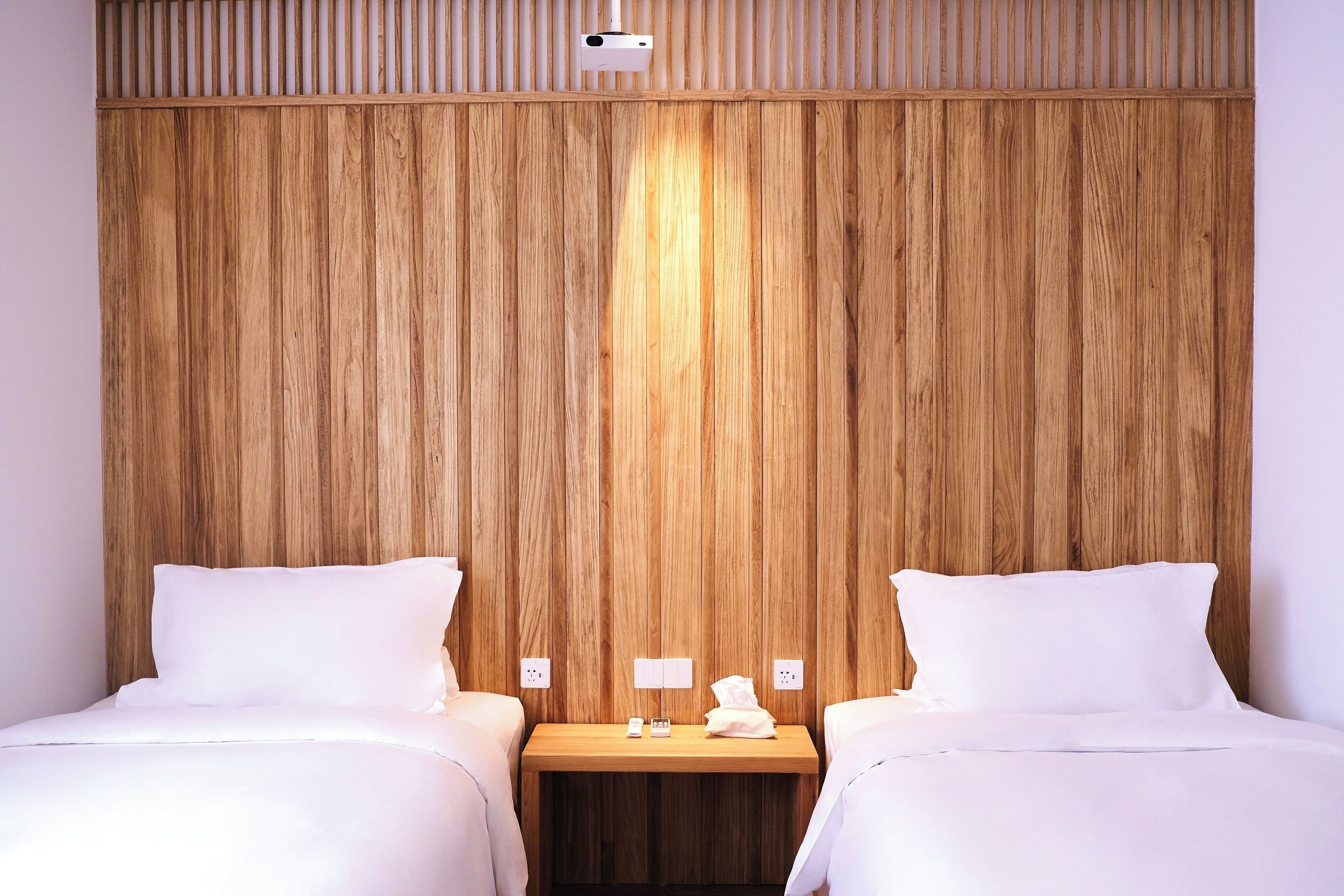 A hotel room with two beds, wooden paneling on the wall, a small wooden nightstand, and a ceiling-mounted projector.