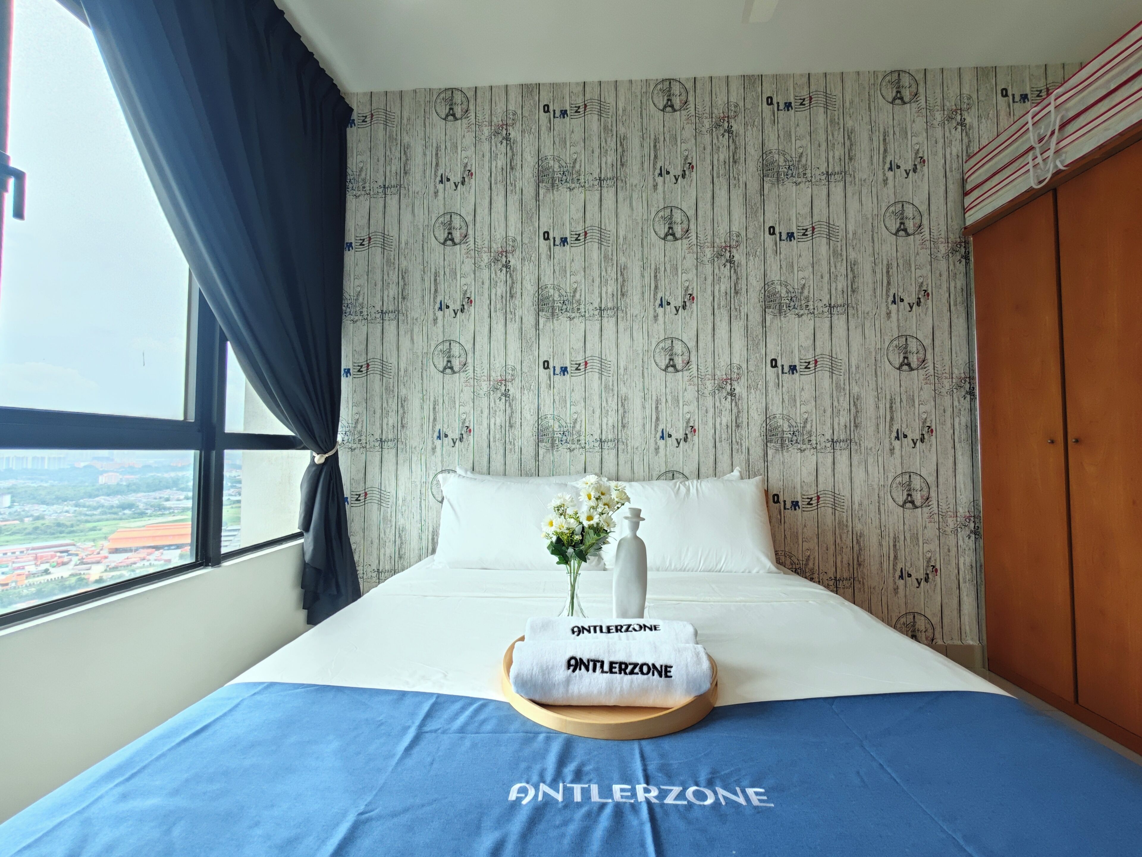 A hotel room with a bed, a window with a view, a curtain, and a wall with a patterned wallpaper.