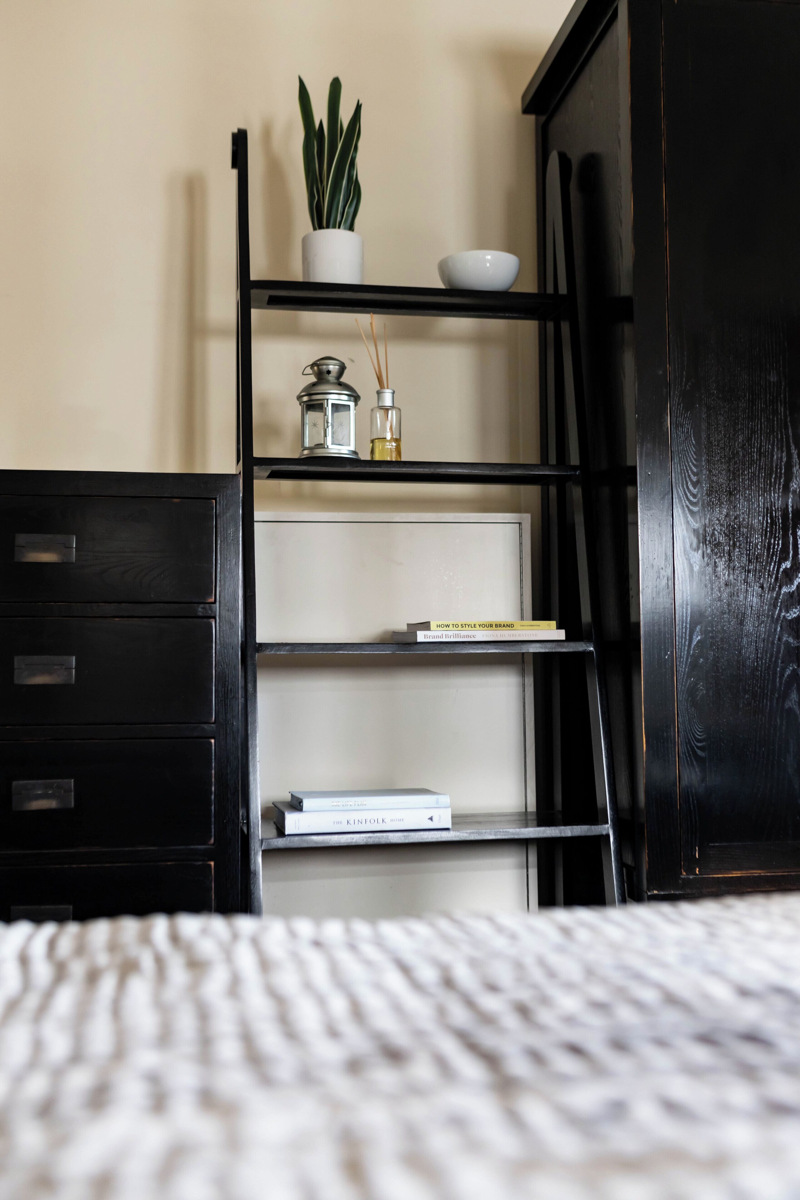 Een slaapkamer met een bed, een zwarte dressoir, een zwarte boekenkast en een potplant.