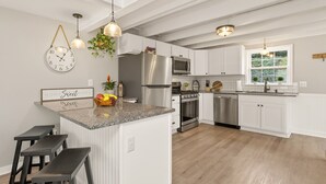 Una cocina moderna con armarios blancos, electrodomésticos de acero inoxidable, una isla central con taburetes, iluminación colgante y un reloj en la pared.