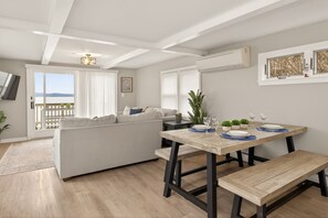 Een moderne woonkamer met een bank, eettafel en een bankje. Er is een balkon met uitzicht, en een airconditioning aan de muur.