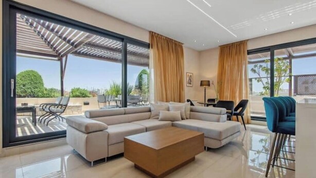 A modern living room with a sectional sofa, a wooden coffee table, and a dining area with a blue chair. Large windows provide a view of the outdoors. - Starry Nights: Penthouse Sauna & Jacuzzi Serenade

 (Glifada)