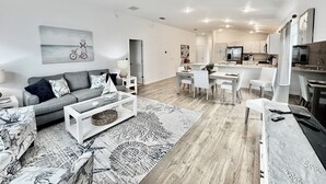 Una sala de estar moderna con un sofá gris, una mesa de centro y un tapete estampado. Un comedor con sillas blancas y una cocina al fondo.