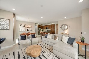 A modern living room with a sectional sofa, a dining area with chairs, a kitchen in the background, and a round wooden coffee table.