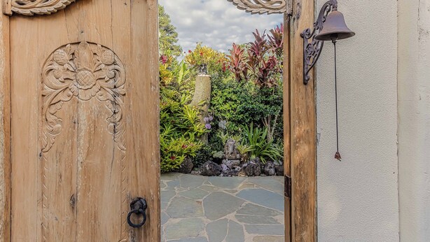 Una puerta de madera con intrincados grabados, un sendero de piedra y un jardín con diversas plantas y una estatua.