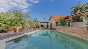 Una piscina con cubierta de madera, un pabellón y una casa con techo de tejas.