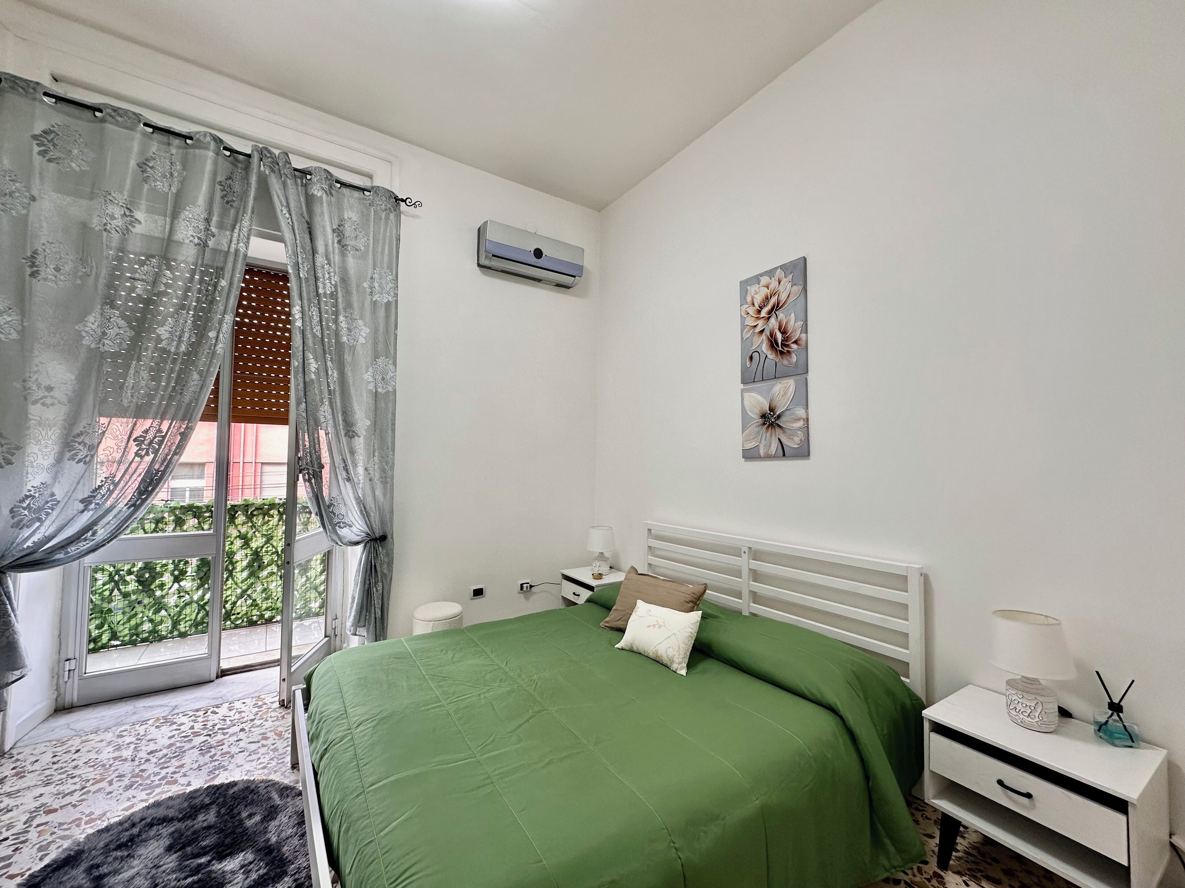 A bedroom with a green bed, a white nightstand, a wall-mounted air conditioner, and a window with a view of greenery.