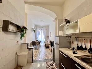 Una cocina estrecha con una zona de comedor al fondo, un refrigerador y diversos utensilios de cocina.