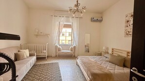 A bedroom with a crib, a sofa, a bed, a chandelier, a window with curtains, an air conditioning unit, and a mirror. - Green Heart House Napoli (Naples)