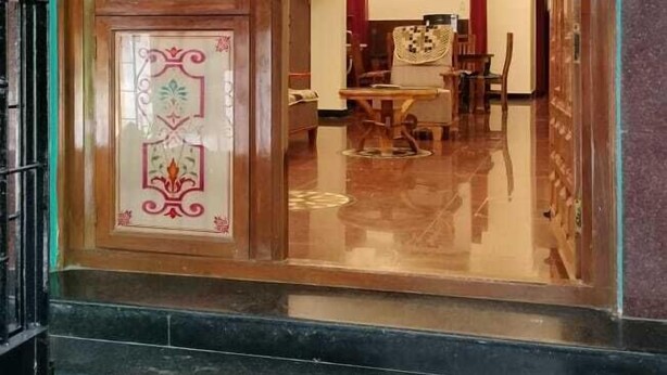 A hotel entrance with a wooden door featuring decorative glass panels. Inside, there is a dining area with a chandelier and a ceiling fan. - Gokulam Villa (Puducherry)
