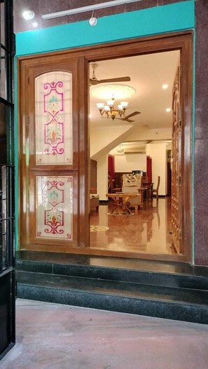 A hotel entrance with a wooden door featuring decorative glass panels. Inside, there is a dining area with a chandelier and a ceiling fan. - Gokulam Villa (Puducherry)