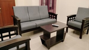 A living room with a grey sofa, two wooden chairs, and a dark wooden coffee table. - Gokulam Villa (Puducherry)
